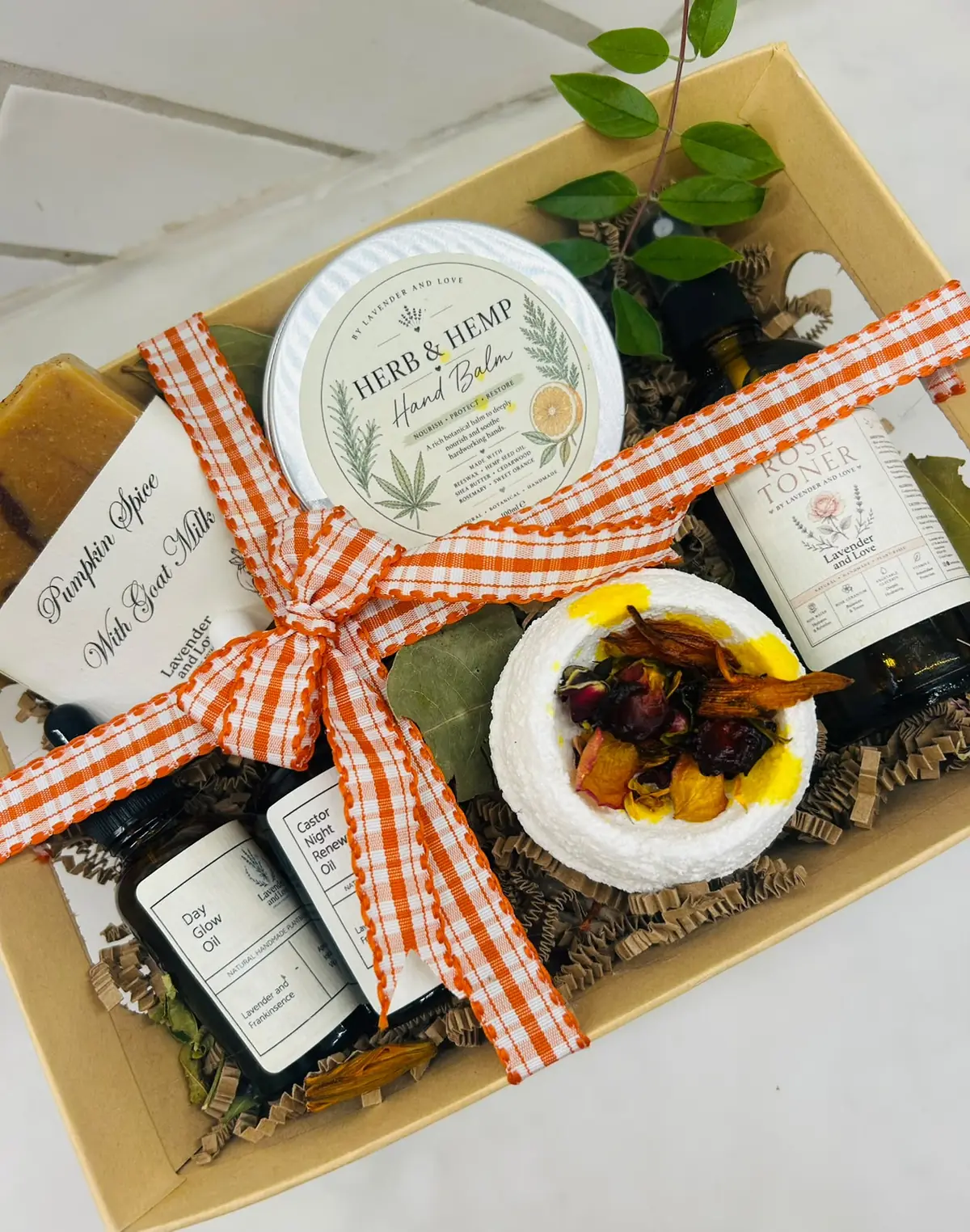 An autumn gift box with Herb and Hemp Hand Balm, Rose Toner, face oils, a shower steamer and pumpkin spice soap, tied with orange gingham ribbon