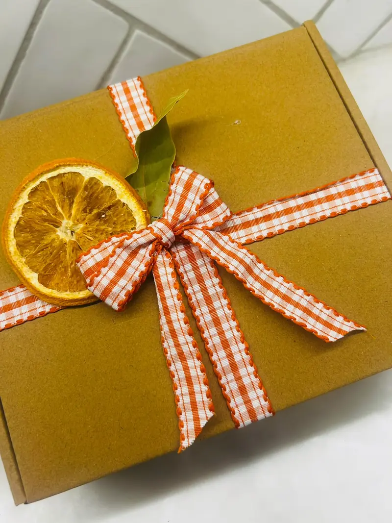 A kraft gift box tied with orange gingham ribbon, a dried orange slice and a bay leaf