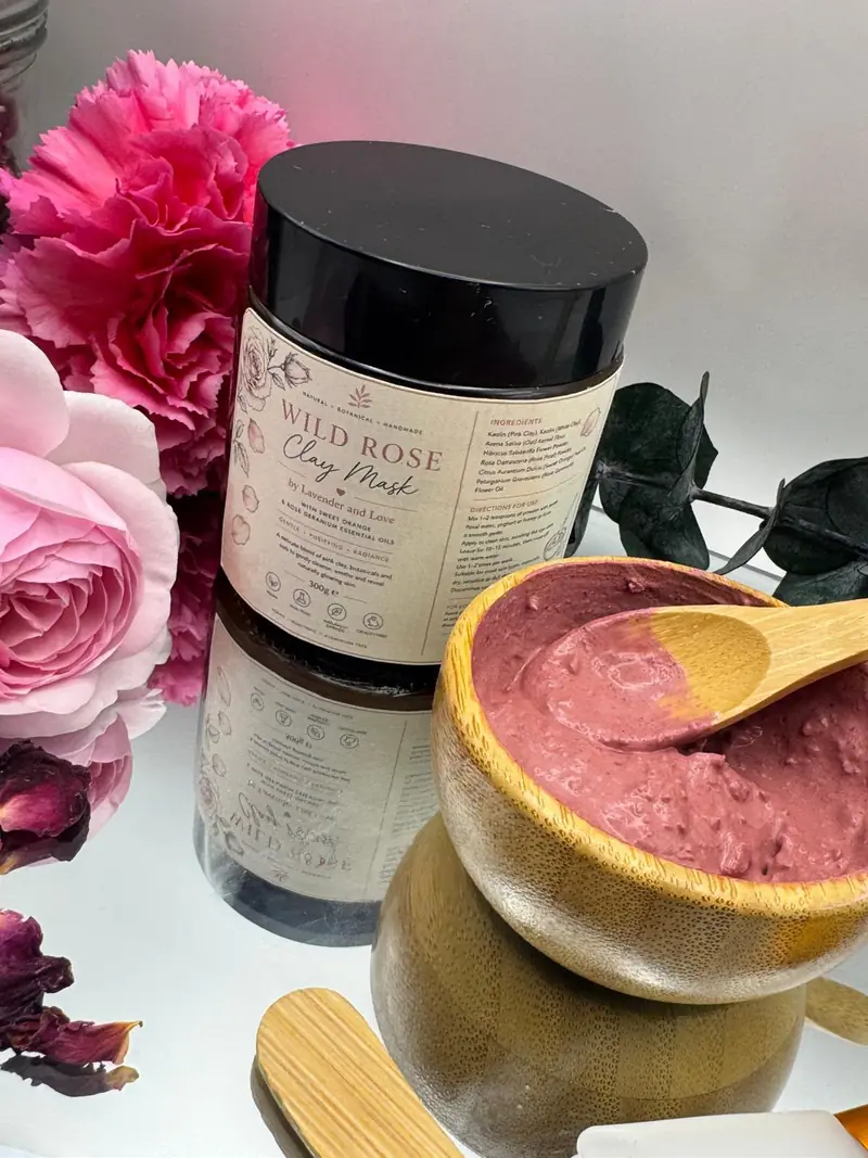 A jar of Wild Rose Clay Mask beside pink roses and carnations, with a bamboo bowl of rose-pink clay
