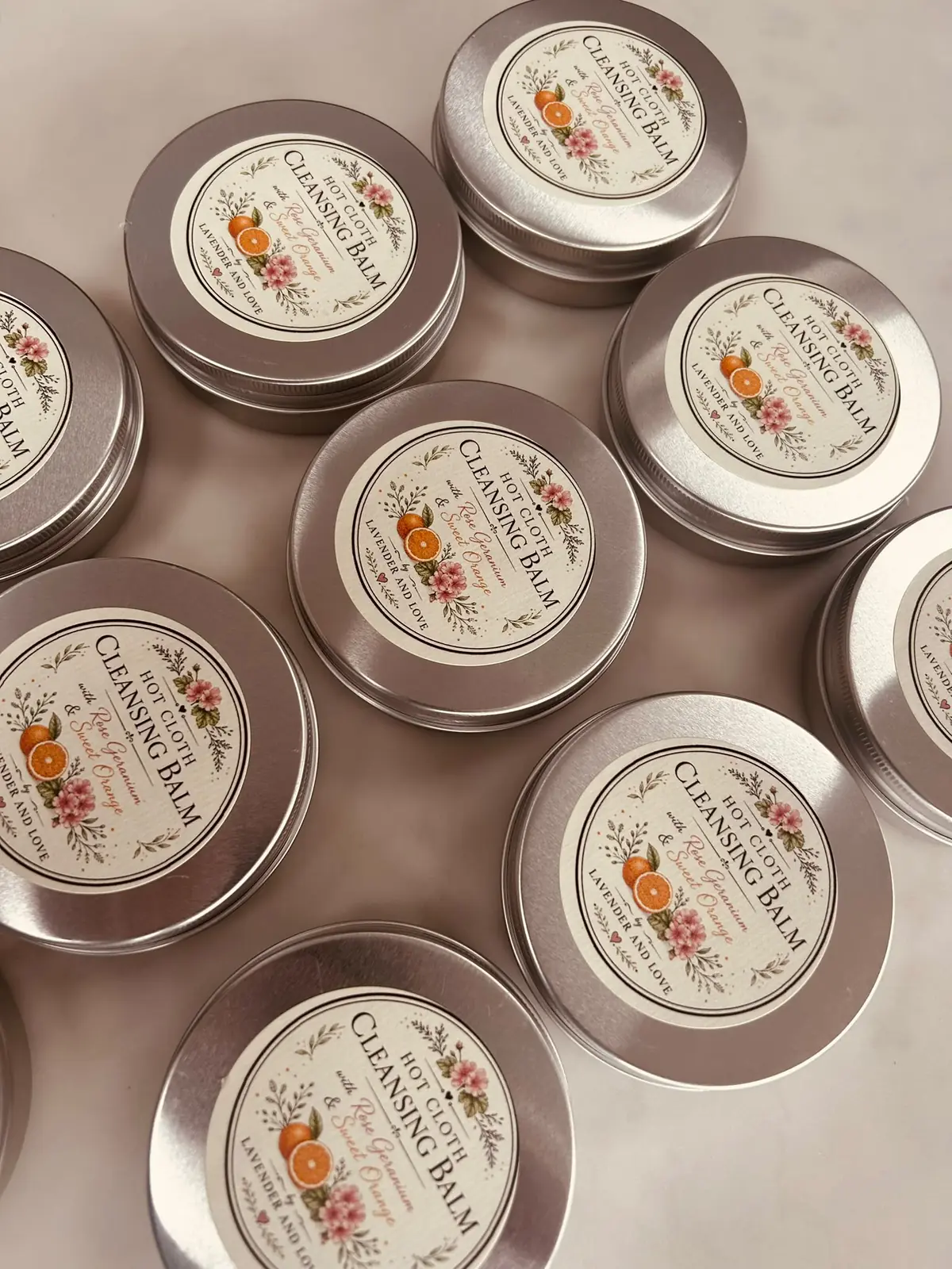 Rows of Hot Cloth Cleansing Balm tins with illustrated rose geranium and orange labels