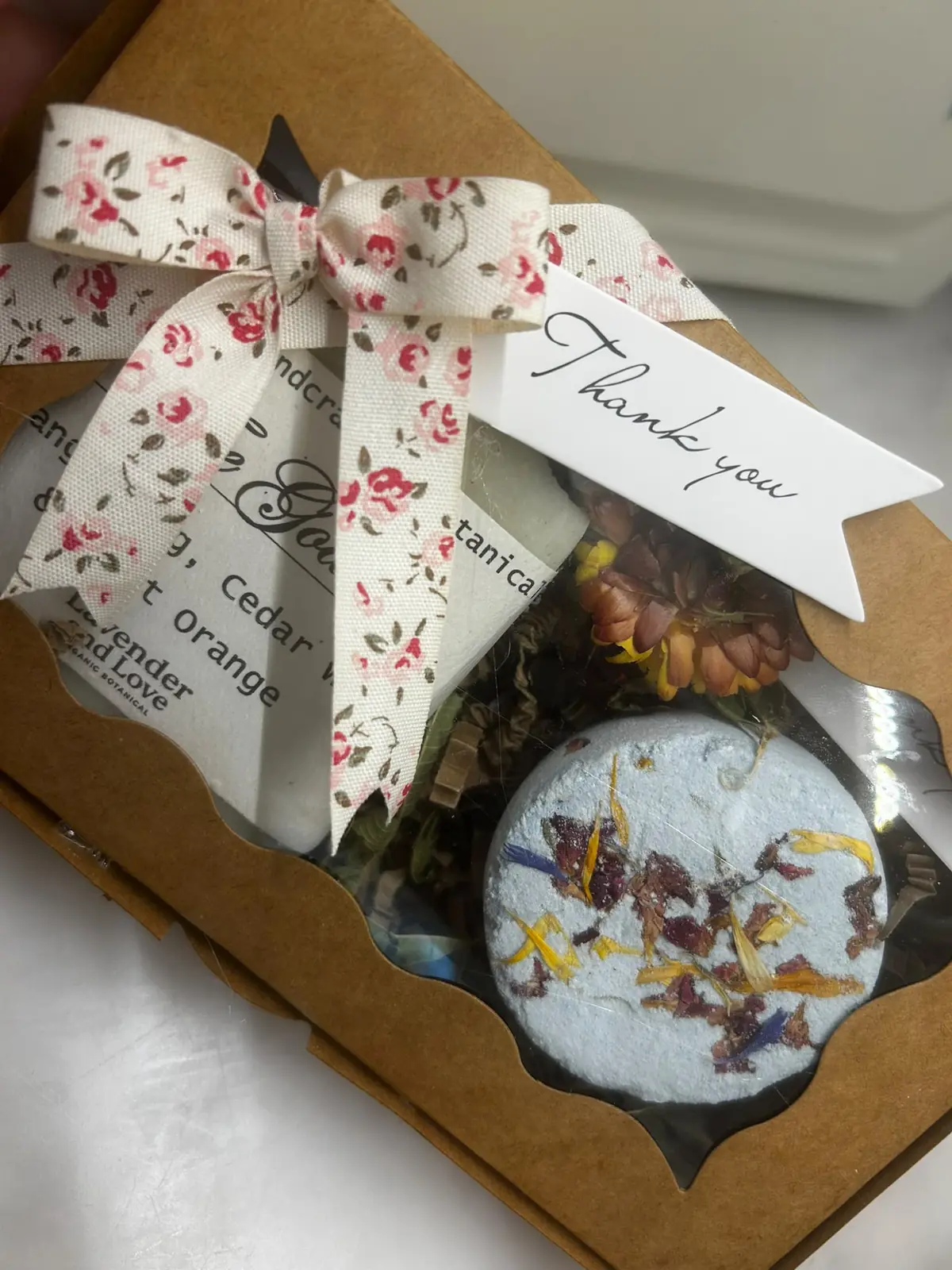 A kraft gift box with a floral ribbon and thank-you tag, holding a handmade soap and a flower-topped shower steamer